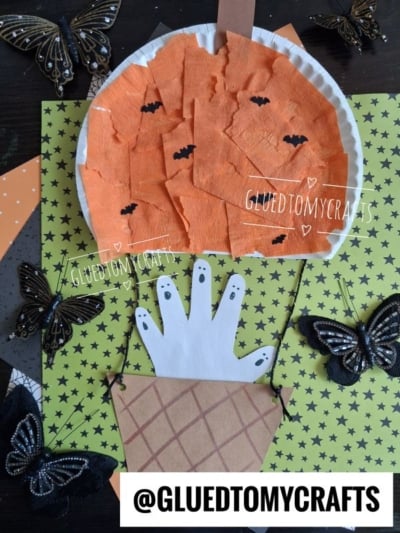 Top-down flat-lay of a kids' Halloween craft with an orange crepe paper pumpkin hot air balloon, handprint ghosts peeking from a paper basket, and decorative black butterflies on a lime-green star-patterned background.