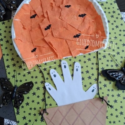 Flat-lay close-up of a paper plate haunted pumpkin hot air balloon Halloween craft, featuring an orange crepe paper pumpkin balloon with black bats, a handprint ghost basket, and glittery black butterfly decorations on a green star-patterned background beside a wooden letter "G."
