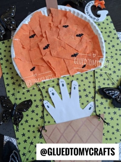 Flat-lay close-up of a paper plate haunted pumpkin hot air balloon Halloween craft, featuring an orange crepe paper pumpkin balloon with black bats, a handprint ghost basket, and glittery black butterfly decorations on a green star-patterned background beside a wooden letter "G."
