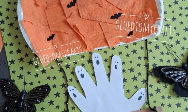 Flat-lay close-up of a paper plate haunted pumpkin hot air balloon Halloween craft, featuring an orange crepe paper pumpkin balloon with black bats, a handprint ghost basket, and glittery black butterfly decorations on a green star-patterned background beside a wooden letter "G."