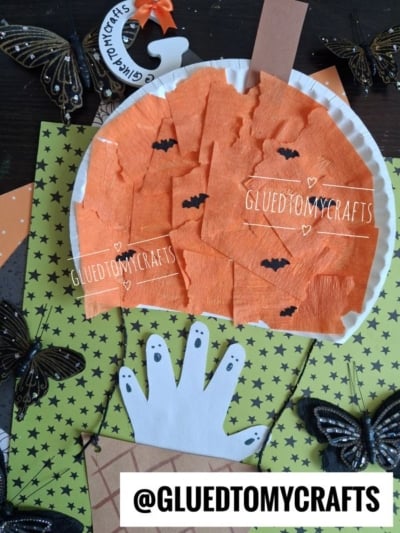 Side angle of a Halloween paper plate craft showing the orange pumpkin balloon connected by black-and-gold metallic twine to a brown woven basket holding five white handprint ghosts.