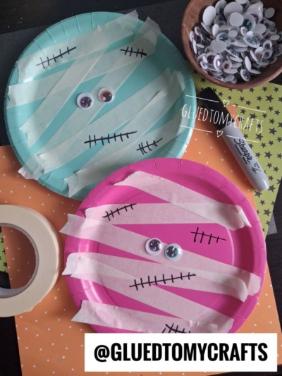 Two paper plates, one green and one pink, decorated as mummies with tape, googly eyes, and marker stitches—making them perfect Halloween Crafts for Kids. Googly eyes, tape, a marker, and orange paper are nearby. @GLUETOMYCRAFTS is written at the bottom.
