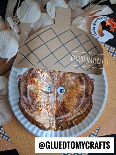 A handmade paint splat acorn craft using a painted paper plate with googly eyes and glitter, topped with a brown paper cap with grid lines, surrounded by leaves. The Instagram handle @GLUEDTOMYCRAFTS is visible.
