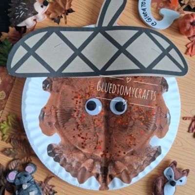 A paper plate acorn craft with googly eyes, brown paint, and glitter, surrounded by artificial leaves, autumn animal decorations, and a GLUEDTOMYCRAFTS label at the bottom.
