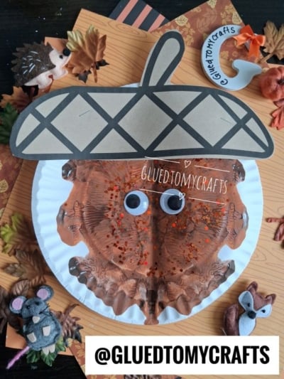 A paper plate acorn craft with googly eyes, brown paint, and glitter, surrounded by artificial leaves, autumn animal decorations, and a GLUEDTOMYCRAFTS label at the bottom.