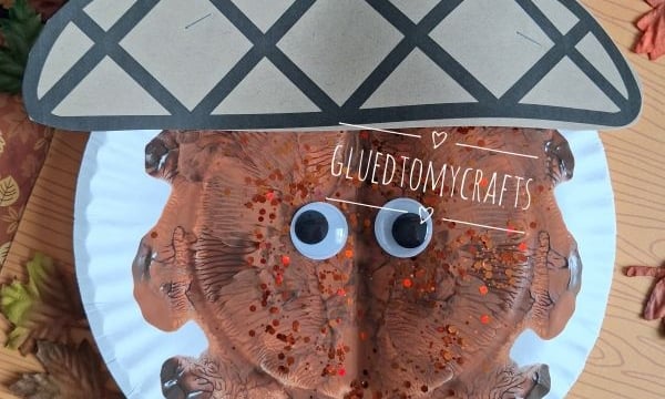 A paper plate acorn craft with googly eyes, brown paint, and glitter, surrounded by artificial leaves, autumn animal decorations, and a GLUEDTOMYCRAFTS label at the bottom.