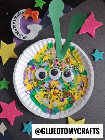 Colorful alien craft on a paper plate with three googly eyes, glitter, and two green antennae. Surrounded by foam stars and a witch hat cutout labeled GluedToMyCrafts..