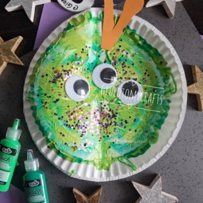 A green paper plate snail craft with large googly eyes, orange antennae, glitter, and paint is surrounded by gold and silver stars and green paint bottles. Tag reads @GLUEDTOMYCRAFTS.