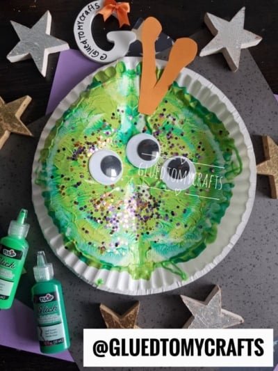 A green paper plate snail craft with large googly eyes, orange antennae, glitter, and paint is surrounded by gold and silver stars and green paint bottles. Tag reads @GLUEDTOMYCRAFTS.