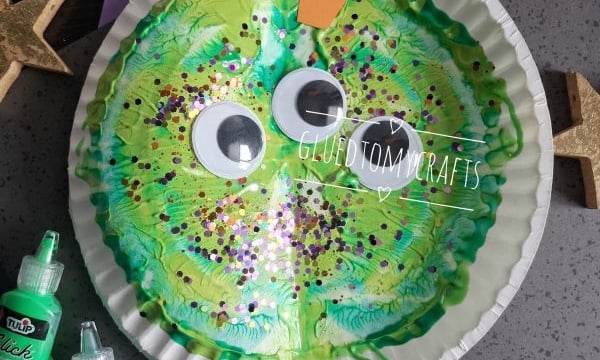 A green paper plate snail craft with large googly eyes, orange antennae, glitter, and paint is surrounded by gold and silver stars and green paint bottles. Tag reads @GLUEDTOMYCRAFTS.