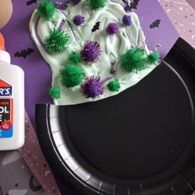 A paper witch hat craft made with green and purple pom-poms, white glue, and purple paper. Black bats decorate the purple background. A bottle of glue sits nearby.