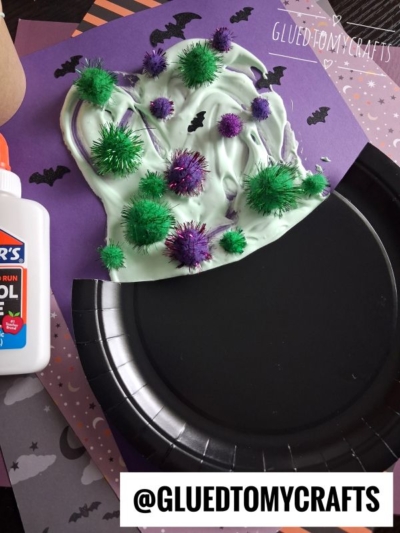 A paper witch hat craft made with green and purple pom-poms, white glue, and purple paper. Black bats decorate the purple background. A bottle of glue sits nearby.