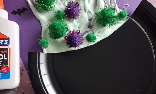 A paper witch hat craft made with green and purple pom-poms, white glue, and purple paper. Black bats decorate the purple background. A bottle of glue sits nearby.