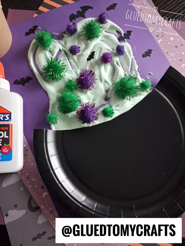A paper witch hat craft made with green and purple pom-poms, white glue, and purple paper. Black bats decorate the purple background. A bottle of glue sits nearby.