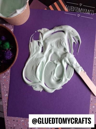A wooden stick spreads light green shaving cream paint on purple paper, surrounded by decorative papers and craft pom-poms. @GLUEDTOMYCRAFTS is written at the bottom.