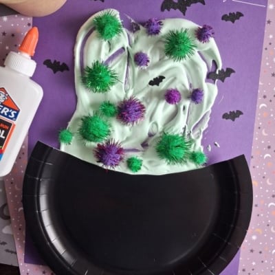 A Halloween craft with a black paper plate cauldron, green slime, glitter pom-poms, and paper bats on a purple background. A glue bottle and cup of green slime are also visible. Text reads @GLUEDTOMYCRAFTS..