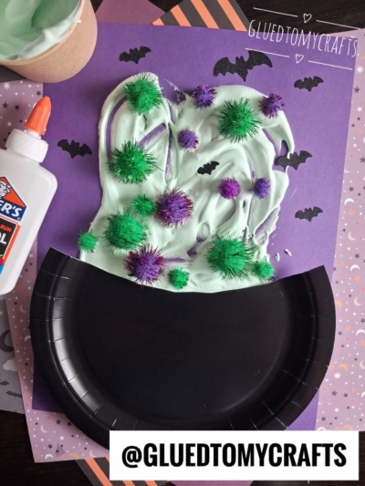 A Halloween craft with a black paper plate cauldron, green slime, glitter pom-poms, and paper bats on a purple background. A glue bottle and cup of green slime are also visible. Text reads @GLUEDTOMYCRAFTS..