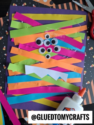 Colorful paper strips and googly eyes are arranged on a purple paper, surrounded by glue, scissors, and other craft materials—perfect for Halloween Crafts for Kids. A white paper strip with zigzag edges is also visible. Text reads @GLUEDTOMYCRAFTS.