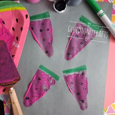 Four painted watermelon slices on gray paper, created using pink paint, black dots, and green marker. Art supplies like paint bottles, a plate, brush, and marker are nearby. @GLUEDTOMYCRAFTS watermark is present.