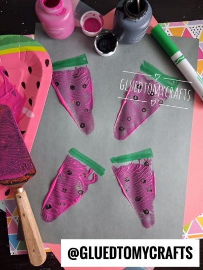 Four painted watermelon slices on gray paper, created using pink paint, black dots, and green marker. Art supplies like paint bottles, a plate, brush, and marker are nearby. @GLUEDTOMYCRAFTS watermark is present.
