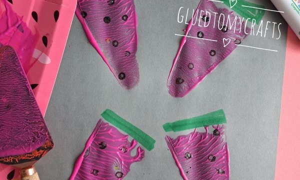 Four painted watermelon slices on gray paper, created using pink paint, black dots, and green marker. Art supplies like paint bottles, a plate, brush, and marker are nearby. @GLUEDTOMYCRAFTS watermark is present.