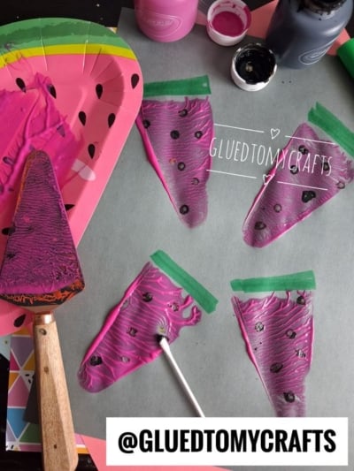 Several painted watermelon slice shapes, some with black seeds, are drying on grey paper with two bottles of paint, a metal spatula, and a disposable watermelon-themed plate nearby. @GLUEDTOMYCRAFTS is written on the image.