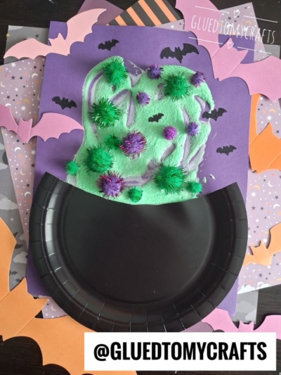 A craft showing a black cauldron created with puffy paint, filled with green, purple, and bumpy potion, surrounded by paper bats on orange and purple paper. @GLUEDTOMYCRAFTS is written on the bottom.