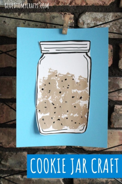 A paper craft of a cookie jar with cookies inside is clipped to a string against a brick wall. Perfect for a cookie jar craft activity, this display could even feature pom pom stamped cookies for added texture. Cookie Jar Craft is written at the bottom in white on a blue background.
