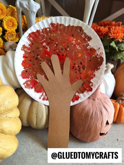 A paper plate painted like fall leaves with a brown paper hand as a tree trunk, surrounded by pumpkins and flowers—making it a perfect addition to your FALL CRAFTS FOR KIDS collection. The @GLUEDTOMYCRAFTS username is visible at the bottom.