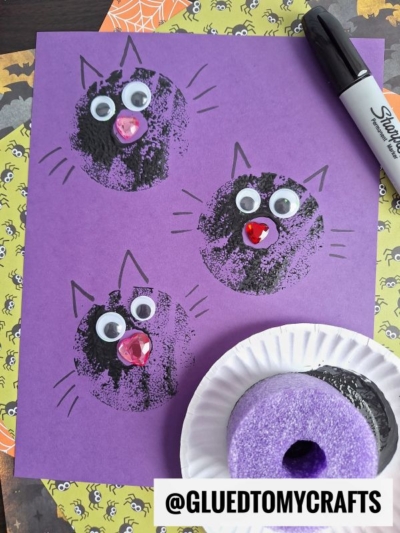 Purple paper with three black-ink stamped cat faces, each decorated with googly eyes, gem noses, and drawn whiskers. A pool noodle stamped cat design is featured alongside a foam stamp and marker. Halloween-themed paper is underneath. @GLUEDTOMYCRAFTS is written at the bottom.