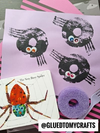 A craft scene with a Eric Carle Busy Spider book, two purple pool noodles, and a purple sheet featuring spider prints with googly eyes. @GLUEDTOMYCRAFTS is labeled at the bottom.