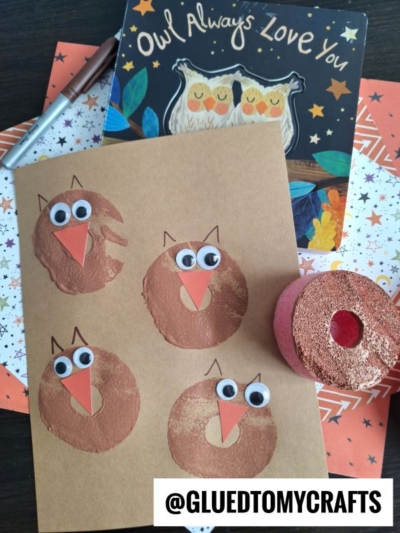 Brown paper with four pool noodle stamped owl crafts made from paint circles, googly eyes, and orange paper beaks. An owl book and patterned papers sit in the background, while a marker and a paint tool are nearby. @GLUEDTOMYCRAFTS text at the bottom.