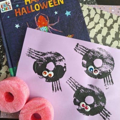 A Halloween craft scene with a Happy Halloween book, ghost-patterned paper, two pink pool noodles, and a pink sheet featuring spider prints with googly eyes. @GLUEDTOMYCRAFTS is labeled at the bottom.