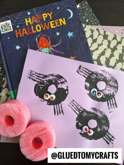 A Halloween craft scene with a Happy Halloween book, ghost-patterned paper, two pink pool noodles, and a pink sheet featuring spider prints with googly eyes. @GLUEDTOMYCRAFTS is labeled at the bottom.