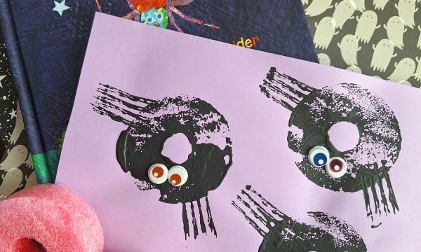 A Halloween craft scene with a Happy Halloween book, ghost-patterned paper, two pink pool noodles, and a pink sheet featuring spider prints with googly eyes. @GLUEDTOMYCRAFTS is labeled at the bottom.