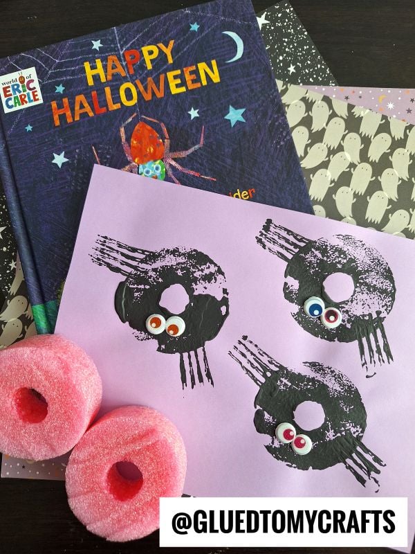 A Halloween craft scene with a Happy Halloween book, ghost-patterned paper, two pink pool noodles, and a pink sheet featuring spider prints with googly eyes. @GLUEDTOMYCRAFTS is labeled at the bottom.