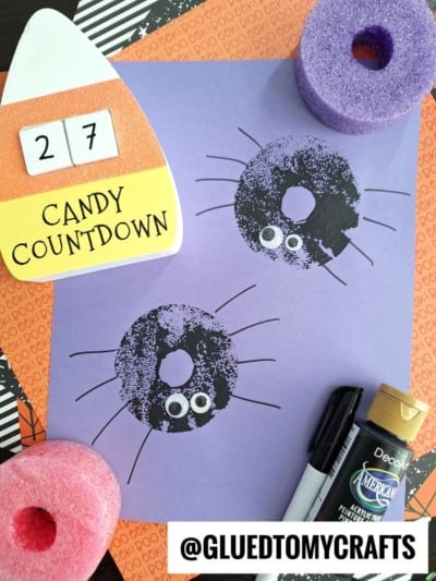 Pool noodle stamping Halloween craft: two black spider prints with googly eyes on purple paper, surrounded by a foam pool noodle ring, black paint, marker, and a candy corn countdown block on an orange “Boo!” background.