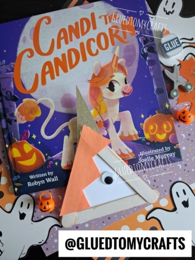 A children’s book called Candi the Candicorn is surrounded by Halloween decorations, perfect for a festive halloween story time. A candy corn craft made from paper, a popsicle stick, and a googly eye is perched on top of the book.