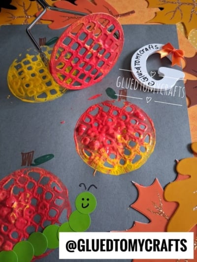 A gray paper with red and yellow pumpkin prints, orange leaves, a green caterpillar sticker, and a craft roller. Text reads @GLUEDTOMYCRAFTS. A small autumn crafts sign with a bow is in the corner.