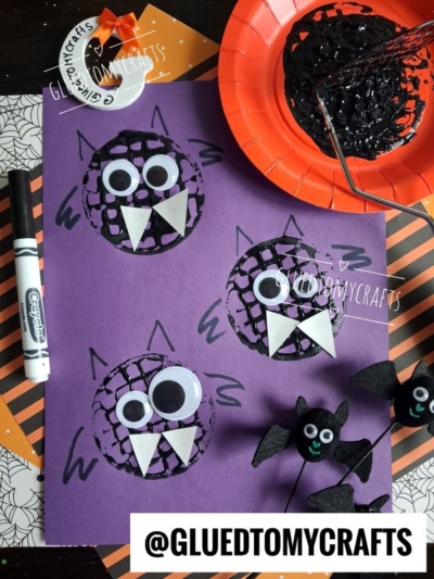 Purple paper features three black doily bats with googly eyes and white fangs, surrounded by craft supplies, a marker, small bat decorations, and hints of a potato masher stamped bat project. The image includes the @GLUEDTOMYCRAFTS watermark.