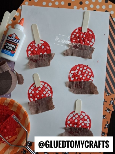 Six paper crafts resembling apple pies with popsicle sticks are glued to white paper. Nearby, a bottle of Elmer’s glue, a paint tray with red paint—perfect for a potato masher stamped craft—and Halloween-themed paper add festive flair. @GLUEDTOMYCRAFTS is written at the bottom.