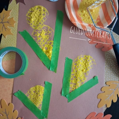 Brown paper with painted yellow corn shapes and green tape for husks, surrounded by paper leaves. A potato masher with yellow paint sits on an orange-striped plate. Craft tape is also visible.