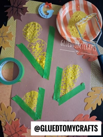 Brown paper with painted yellow corn shapes and green tape for husks, surrounded by paper leaves. A potato masher with yellow paint sits on an orange-striped plate. Craft tape is also visible.