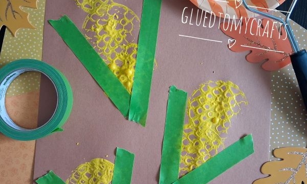 Brown paper with painted yellow corn shapes and green tape for husks, surrounded by paper leaves. A potato masher with yellow paint sits on an orange-striped plate. Craft tape is also visible.