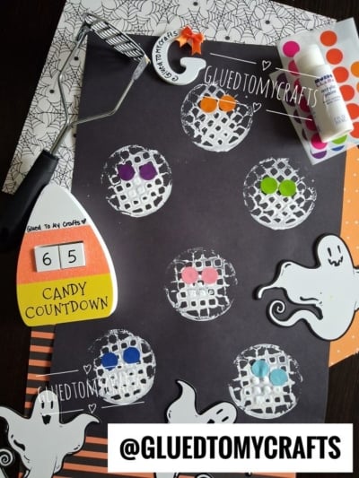 Kids’ Halloween craft showing six white potato-masher stamped circles on black paper, each decorated with colorful sticker eyes, surrounded by paint, ghost cutouts, and festive craft supplies.