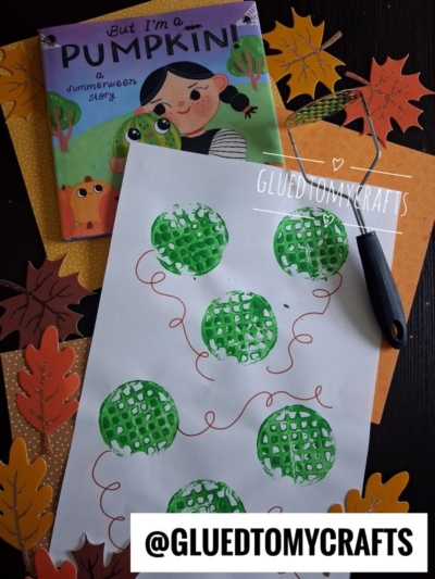 A children’s book, fall leaves, and a pumpkin-themed craft made with green paint and bubble wrap are on a dark table. A paint roller, a potato masher stamped craft, and the @GLUEDTOMYCRAFTS label are also visible.