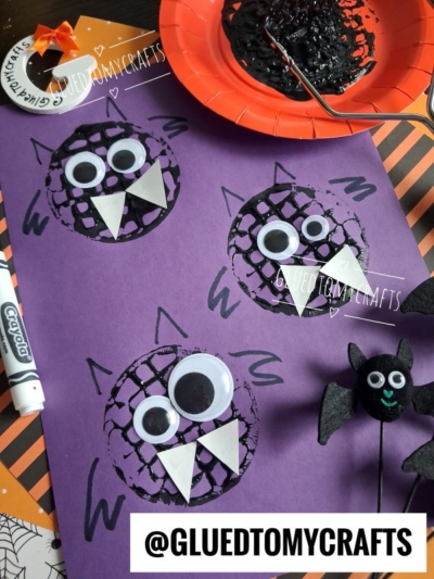 Purple paper with three black spiders made from drawn circles, large googly eyes, and white triangle teeth. Craft supplies, a black marker, and a potato masher stamped bat are nearby. @GLUEDTOMYCRAFTS appears at the bottom.