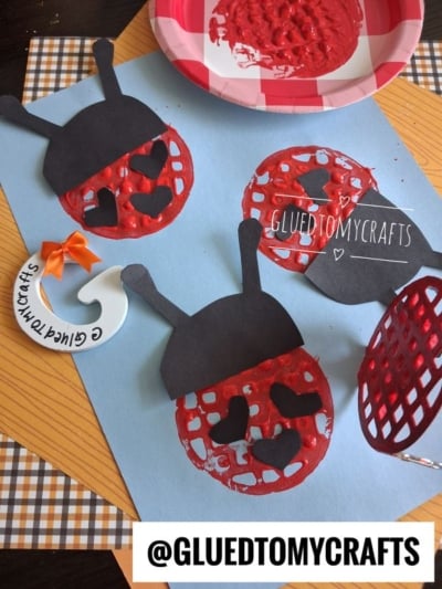 A blue paper displays ladybug art made from red paint circles and black paper. Paint supplies, a paper plate with red paint, and a waffle spatula are arranged around the artwork. @GLUEDTOMYCRAFTS is visible.