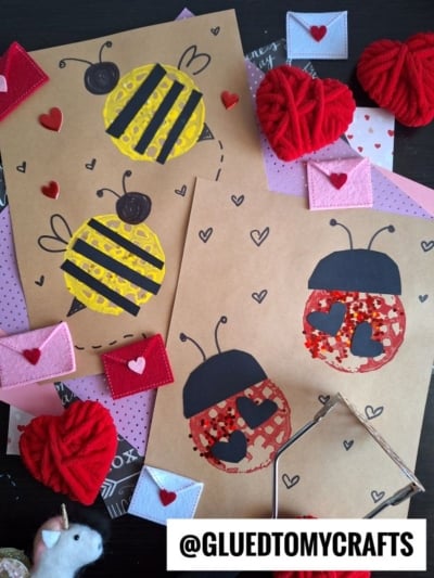 Two handmade cards on a table, each with a bee or ladybug made from colored paper and confetti. Red felt hearts, pink envelopes, and yarn hearts decorate the scene. @GLUEDTOMYCRAFTS is written at the bottom.