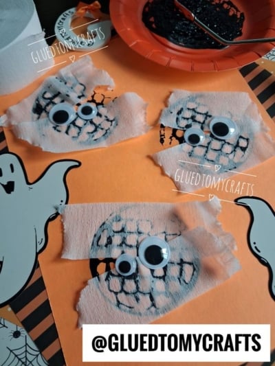 Googly-eyed mummy faces made from black mesh and white crepe paper are displayed on orange paper with ghost cutouts. A red plate with black paint and a pumpkin sticker are in the background.